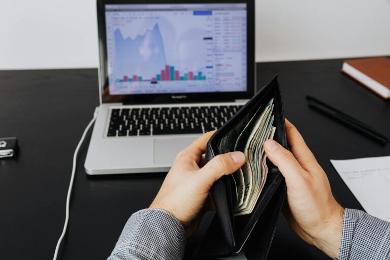 Man holding a leather wallet with bills in front of a laptop displaying financial graphs.